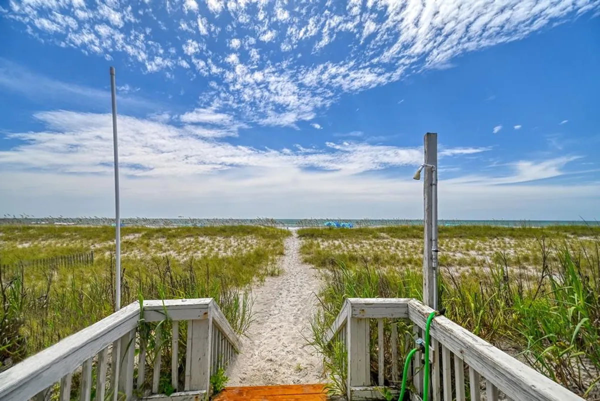 A serene beach pathway leads visitors toward the vibrant blue ocean beneath a cloud-filled sky. The image captures the inviting nature of the shoreline, with suggestions of sunbathing, sandcastle building, and gentle waves ready for swimming.
