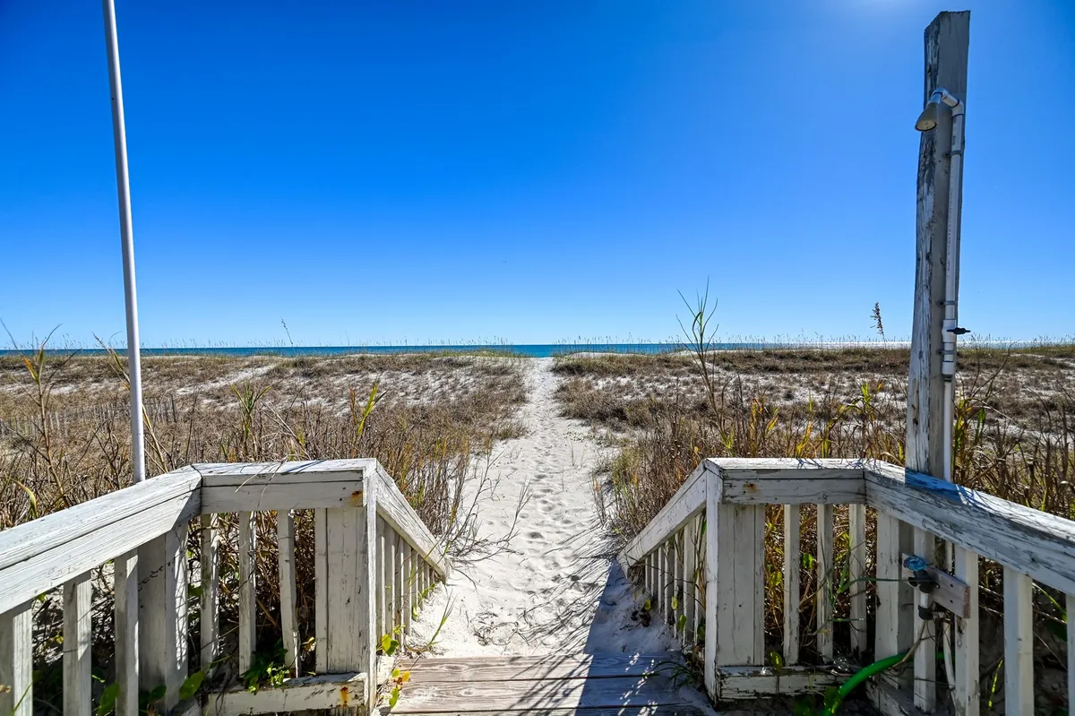 Wooden steps and a boardwalk path leading over grassy dunes directly to the sandy beach, with the ocean horizon in the background. The scene is framed by natural grasses and subtle dunes, evoking a sense of tranquil transition from house to shore.