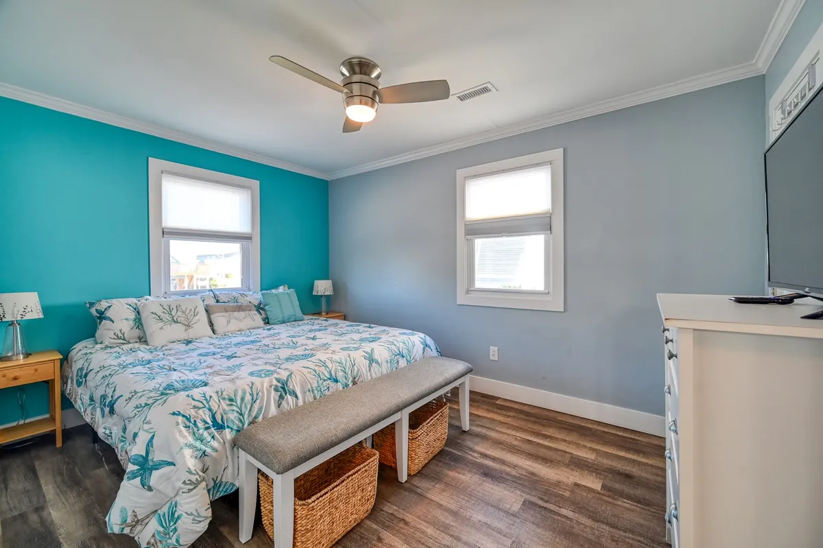 A tranquil bedroom featuring a bed with floral bedding, a turquoise accent wall, ceiling fan, and a wooden bench with storage. Natural light pours in, creating a calm and serene environment, ideal for rest and relaxation.