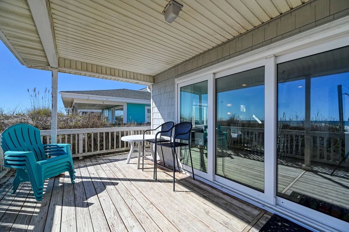A tranquil exterior shot featuring a blue Adirondack chair and other seating on the deck, with glass sliding doors leading to the house. The area overlooks scenic views and is ideal for relaxing, socializing, or watching the beach sunsets after a day filled with pet-friendly fun.