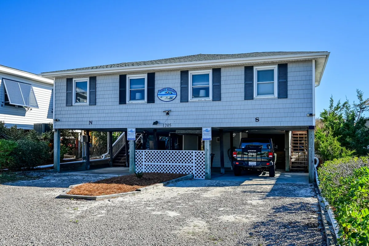This exterior image showcases SeaClusion House bathed in sunlight, with a welcoming driveway, well-maintained landscaping, and inviting outdoor spaces. The house appears approachable and family-friendly, offering plenty of room for outdoor activities—perfect for guests arriving with pets in tow.