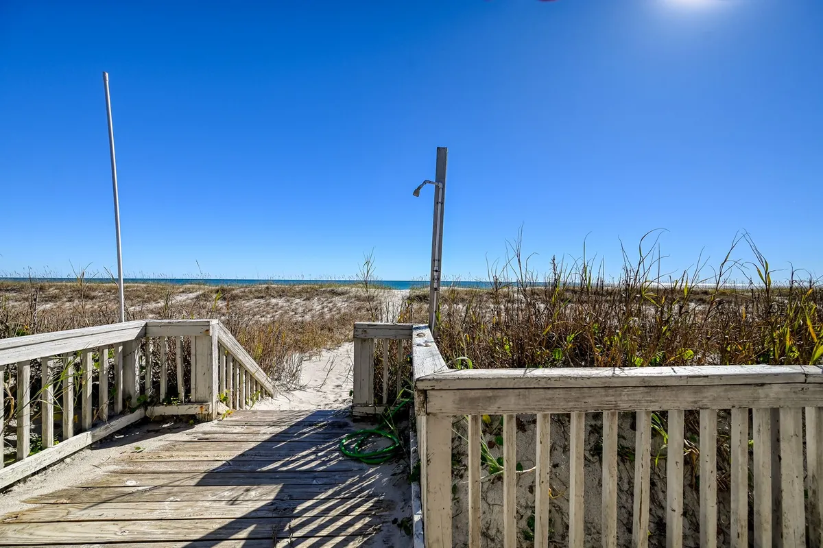 A wooden walkway extends from the house through tall beach grass, leading directly to the sandy shore. The expansive ocean view stretches out beyond the grasses, creating an inviting path for beachgoers. The scene is tranquil, with soft natural light and a sense of peaceful seclusion.